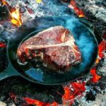 t bone steak being cooked on a cast iron pan outdoors